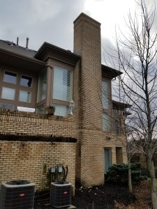 Chimney Crowns Repair 10 A two-story brick house with large windows and a tall chimney. There are two air conditioning units and utility meters outside, along with a bare tree and some small bushes. The sky is overcast.