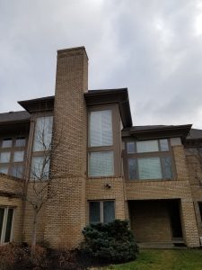 Chimney Crowns Repair 11 A two-story brick building with large windows featuring white shutters, a tall chimney, and a covered entrance. Leafless trees and bushes are in the front yard under a cloudy sky.