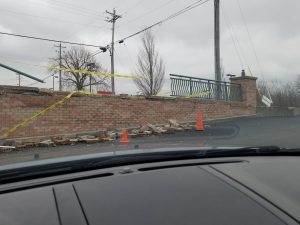 A damaged brick wall with yellow caution tape and orange cones blocks off debris on the ground. The photo is taken from inside a car on a cloudy day. Power lines and leafless trees are visible in the background.
