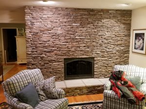 A living room with a stone fireplace, two plaid armchairs with cushions, a jacket draped over one chair, and a framed photo on the wall. The floor is wooden with a patterned rug.