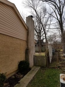 Chimney Crowns Repair 4 A brick house with a large, weathered chimney on its side, a narrow concrete sidewalk, small bushes, a wooden gate, trees without leaves, and neighboring houses in the background on an overcast day.