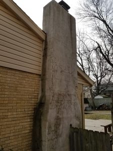 A tall, cracked concrete chimney attached to a yellow brick house, showing extensive surface damage. Leafless trees and a wooden fence are visible nearby, with a cloudy sky in the background.