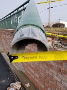 A large green metal pole lies broken and detached from a brick wall, with yellow Premier Plus caution tape in front of the scene. Pieces of brick are scattered on the ground nearby.