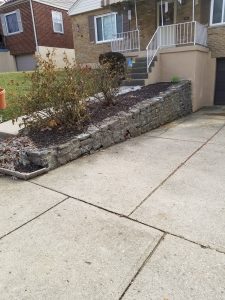 A concrete driveway leads up to a stone retaining wall with a small garden bed and some shrubs in front of a brick house with white railings on the porch. A brown house is visible to the left.