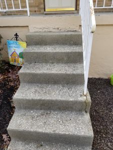 Masonry And Stonework 7 Six concrete steps with a white metal railing lead up to a door. There is a crack on the bottom step, and a colorful garden flag to the left reads Grandkids Loved Here. Brown mulch and leaves surround the steps.