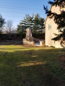 A partially built outdoor fireplace or brick structure stands on a concrete pad in a grassy backyard, next to a brick house and surrounded by trees under a clear blue sky.