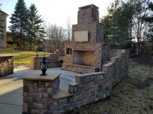 Outdoor stone fireplace with two lanterns, surrounded by a low stone wall, set on a concrete patio. There are trees and a grassy yard in the background, with sunlight filtering through the branches.