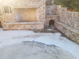 Outdoor stone fireplace with a built-in bench and a small arched tunnel underneath. Water is pooled and spreading on the concrete in front of the structure, with some wet patches and cracks visible.