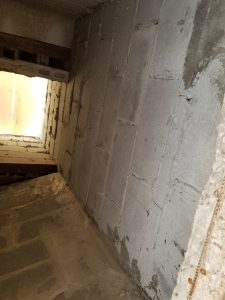 A view looking up inside a narrow shaft or chimney constructed from gray concrete blocks, with light coming in from an opening at the top. The walls appear rough and unfinished.
