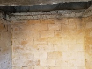 A close-up view of a corner where two beige concrete block walls meet, with a rough, unfinished concrete ceiling above. The walls show stains and wear.
