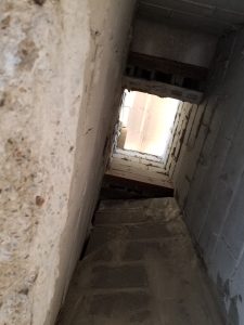 View looking up inside a narrow, unfinished concrete chute or shaft with rough brick and mortar walls, exposed beams, and sunlight visible through an opening at the top.
