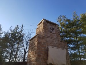 A tall, stone outdoor structure with a covered opening and a plaque featuring the letter P. Green trees and a clear blue sky are in the background.