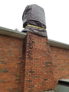 Chimney Crowns Repair 6 A brick chimney extends from the roof of a house, with the top covered by a large, brown tarp secured with yellow tape. Dark stains run down the brick below the covered area.