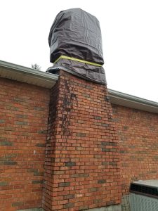 A red brick chimney extends from the side of a house, with its top wrapped in a large gray tarp secured by a yellow strap. Black stains are visible on the bricks, and an HVAC unit sits nearby.
