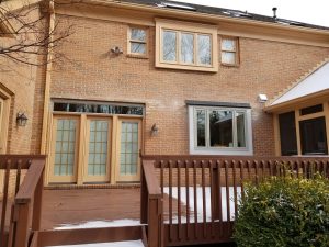 A brick house with tan trim features French doors, a large window, and a wooden deck partially covered with snow. Bushes and a small amount of snow are visible in the foreground.