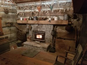 A rustic log cabin room with a stone fireplace, burning wood stove, and various antique tools. Animal skulls and antlers decorate the log walls, and metal kettles and pots sit on the wooden mantel.