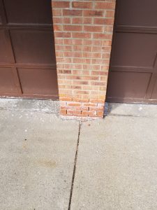 A brick pillar in front of a brown garage door shows signs of water damage and white salt deposits at the base, with scattered debris on the surrounding concrete sidewalk.