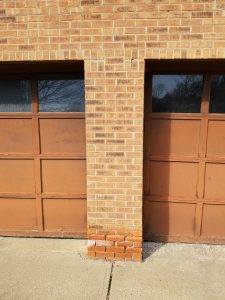 A brick column separates two brown garage doors. The bottom row of bricks on the column is a noticeably different color, appearing more orange than the others. The concrete driveway is in the foreground.