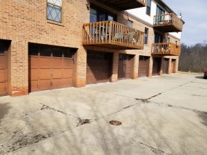 A row of brown garage doors beneath brick apartment balconies, with a large cracked concrete driveway in front and a car partially visible on the far right.