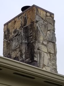 A close-up view of a stone chimney with a metal cap, rising above a beige roofline against a cloudy sky.