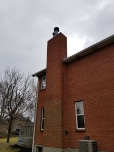 A brick chimney attached to the side of a red-brick house extends above the roofline under a cloudy sky. Leafless trees and a wooden deck are visible in the background.