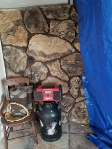 Masonry And Stonework 4 A rustic corner with a stone wall, a wooden chair holding a basket and a bag, a large black container with white designs, a red radio on top, and a blue tarp hanging to the right.