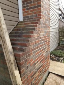 A brick chimney appears to be bulging outward from a house exterior, next to wood planks leaning against the wall. Siding and a small garden area are also visible.