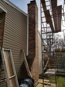 Scaffolding and ladders are set up next to a brick chimney attached to a house with beige siding, indicating ongoing construction or repair work. Leafless trees are visible in the background.