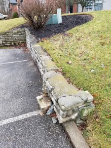 A damaged stone retaining wall by a grassy slope; several stones at the corner are broken and displaced, next to a parking lot with asphalt and a concrete curb.