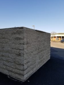 A large beige brick structure stands in a parking lot under a clear blue sky, with a strip mall and cars visible in the background.