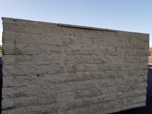 A beige brick wall stands outdoors under a clear blue sky, with sunlight casting shadows on the textured surface. The wall is freestanding and the ground around it is paved with asphalt.