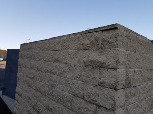 A close-up view of the corner of a beige stone block wall under a clear blue sky, with part of a building and a streetlamp visible in the background.