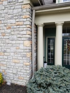 Masonry And Stonework 5 A beige downspout runs along the corner of a stone wall next to two large columns at the entrance of a building, with a green shrub in the foreground and glass doors in the background.