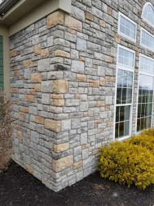 Masonry And Stonework 6 Corner view of a house exterior with light gray and tan stone veneer siding, large white-trimmed windows, yellow-green shrubs, and dark mulch at the base.