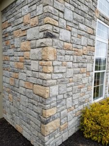A close-up view of the corner of a building with a stone veneer exterior, featuring gray and beige stones. There are white-framed windows on one side and a yellow-green shrub at the base.