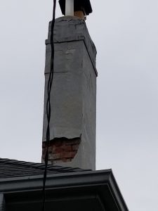 Chimney Repair 11 A damaged chimney with cracked and missing plaster reveals red bricks underneath, situated on the roof of a house against a cloudy sky. A black cable hangs in front of the chimney.