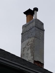 A weathered brick chimney with peeling white paint stands on a roof, topped with a black metal cap and a smaller metal pipe. The sky in the background is overcast.
