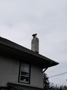 Chimney Repair 12 A white house with a tall, weathered chimney extends above the roofline under a cloudy sky. The chimney has a metal cap and a vent pipe. A window is visible on the houses side.