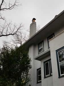 A gray house with a tall chimney and dark-trimmed windows is shown from below, against a cloudy sky. Leafless tree branches frame the upper left corner, and evergreen bushes are in the foreground.