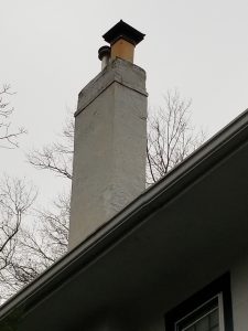 A tall, weathered chimney with a cap rises above a white house roof; bare tree branches are visible in the background against a cloudy gray sky.