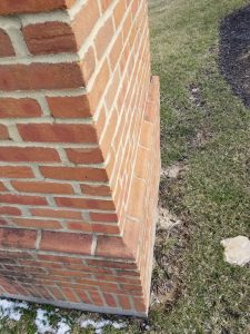 A close-up view of a brick corner structure with layered brickwork, bordered by grass and a small patch of soil. Some snow is visible in the lower left corner.
