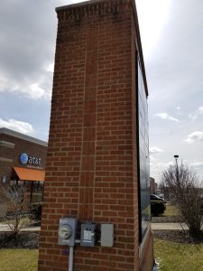A tall brick sign structure stands by a sidewalk with electrical panels attached. In the background is an AT&T Authorized Retailer store with an orange awning. The sky is partly cloudy. Bushes and a parking lot are visible.