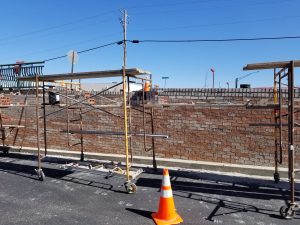 Construction site with brick wall, scaffolding, and orange traffic cone on asphalt. Bricks and tools are visible, and the sky is clear and blue in the background.