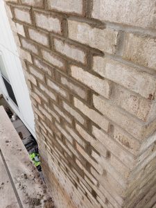 A close-up view of a beige brick wall corner, looking downward from above. The mortar between the bricks is visible, and a person in a green vest is partially seen at the bottom near construction materials.