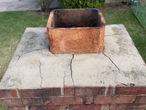 Chimney Crowns Repair 13 A damaged brick chimney with a cracked concrete crown and worn, discolored flue liner, shown from above; grass and a sidewalk are visible in the background.