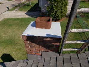 Chimney Crowns Repair 15 A brick chimney with a brown chimney cap is seen from above on a sunny day. The top is freshly coated with white sealant, and a ladder is positioned next to it, resting on a shingled roof.