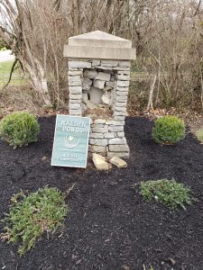 A damaged stone pillar stands in a mulched area with bushes around it. A green sign reading Walden Ponds: A Golf Community leans against the pillar. Trees and grass are visible in the background.