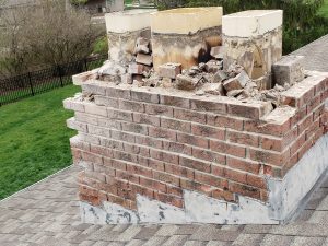 A severely damaged brick chimney on a rooftop with crumbling bricks, missing mortar, and broken sections, exposing the inner structure. Green grass and trees are visible in the background.