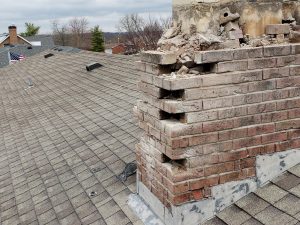 Chimney Crowns Repair 17 A damaged brick chimney with crumbling and missing bricks sits on a shingled roof; debris is scattered around, and neighboring rooftops are visible in the background.