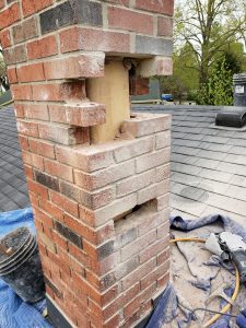 Chimney Repair 9 A brick chimney on a roof with two large rectangular sections of bricks removed, exposing wood inside. Tools, a bucket, and loose bricks are on a blue tarp nearby. Trees are visible in the background.